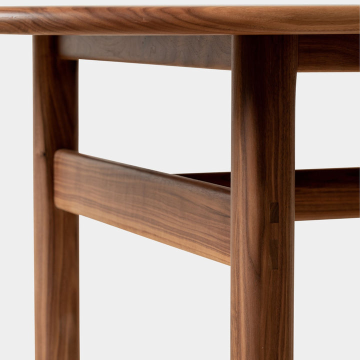 The PALANG Dining Table by Earl Home boasts a robust oval pill shape with bullnose edge design in rich walnut, supported by four legs and a crossbeam. It is available in white oak, maple, or ebonized oak.  This table comes in customizable lengths and top shapes including rectangular or live edge for perfect fitting in any space.