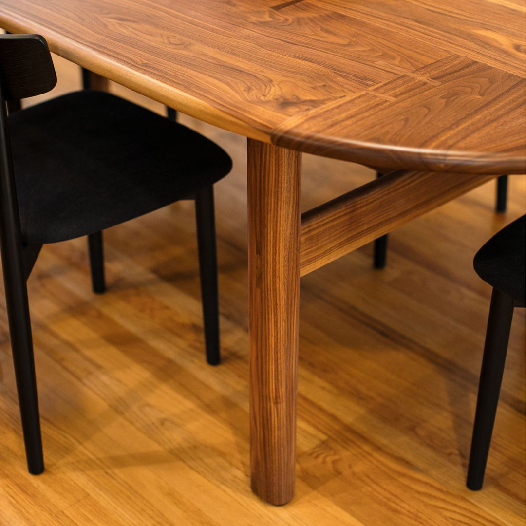 The PALANG Dining Table by Earl Home boasts a robust oval pill shape with bullnose edge design in rich walnut, supported by four legs and a crossbeam. It is available in white oak, maple, or ebonized oak.  This table comes in customizable lengths and top shapes including rectangular or live edge for perfect fitting in any space.