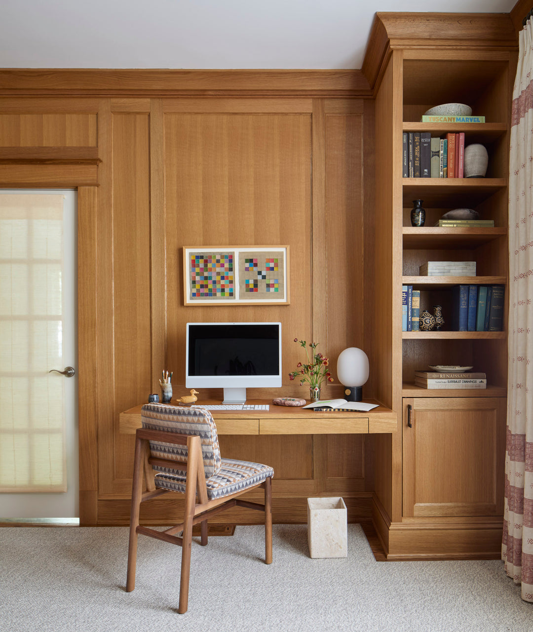 A wooden home office with a computer on a floating desk. A chair is in front, with a bookshelf and decorative items to the right. A door with a blind is on the left. Art is above the desk.