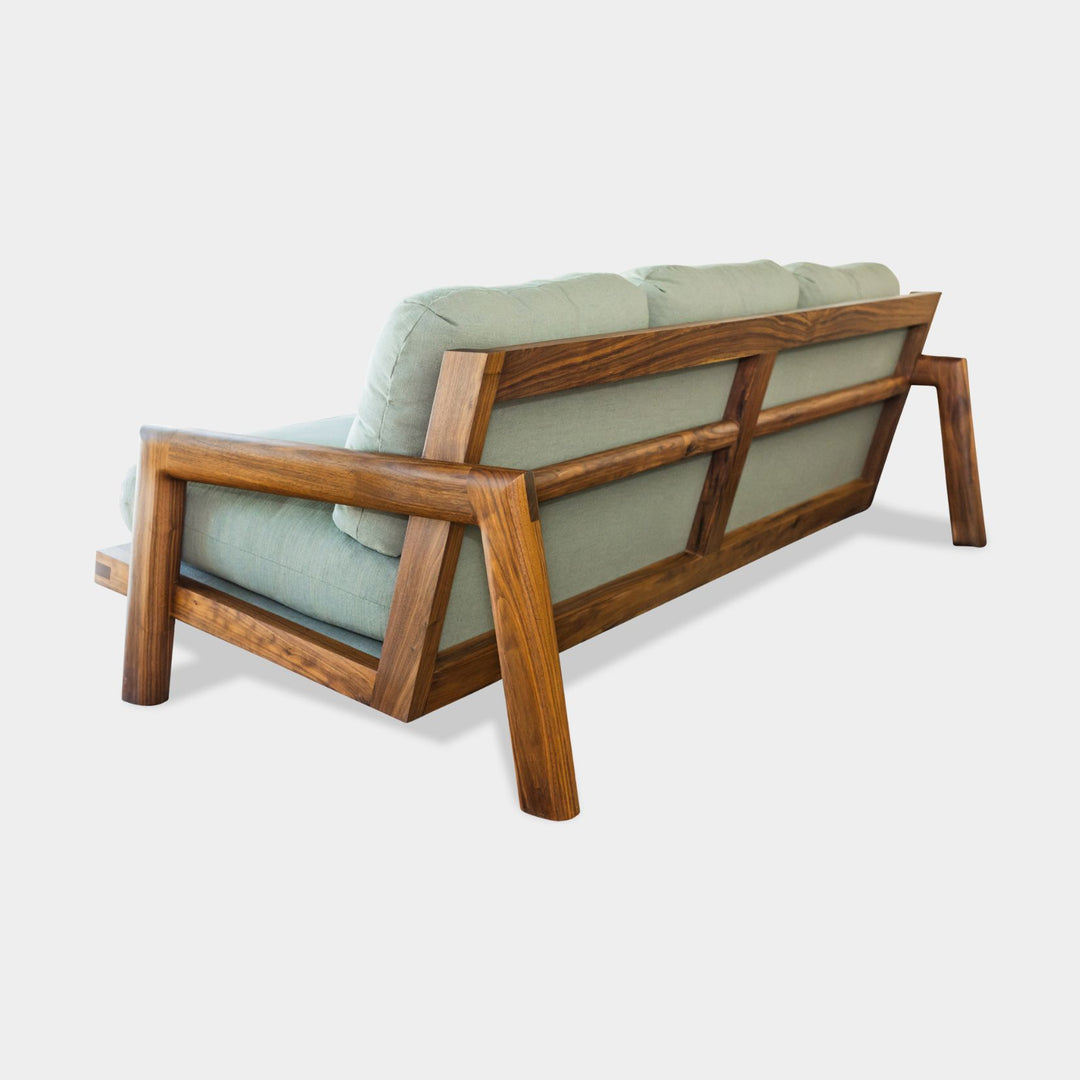 Earl Home's TAMBU Sofa features an American walnut wooden frame with green cushions and three backrests against a minimalist background. Handmade to order, it seamlessly blends elegance with bespoke craftsmanship in its striking design.