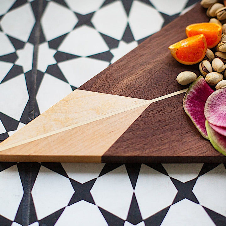 The TRIANGLE Cheese Board by Earl Home is a stylish centerpiece with a triangular design, lighter wood apex, darker base, brass and wood accents, and a central vertical division.