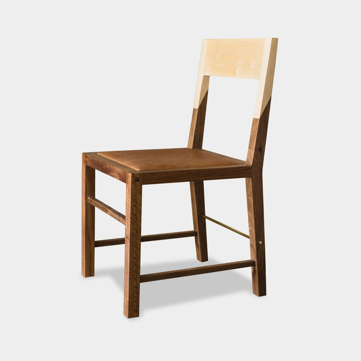 The TUPELA Chair by Earl Home features a minimalist, modern design with a light maple backrest and walnut solid wood seat and frame. Its custom camel leather upholstered seat enhances comfort and style, perfectly blending elegance with craftsmanship.