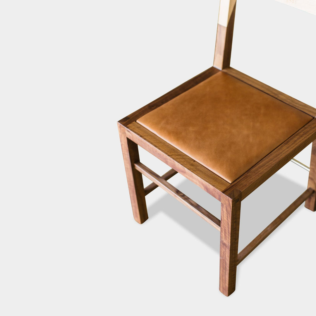 The TUPELA Chair by Earl Home features a minimalist, modern design with a light maple backrest and walnut solid wood seat and frame. Its custom camel leather upholstered seat enhances comfort and style, perfectly blending elegance with craftsmanship.