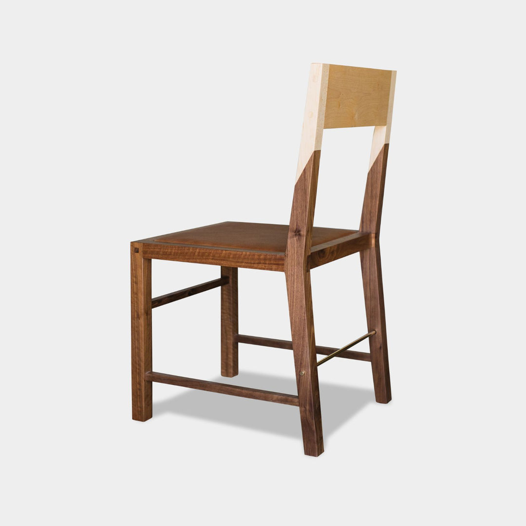 The TUPELA Chair by Earl Home features a minimalist, modern design with a light maple backrest and walnut solid wood seat and frame. Its custom camel leather upholstered seat enhances comfort and style, perfectly blending elegance with craftsmanship.