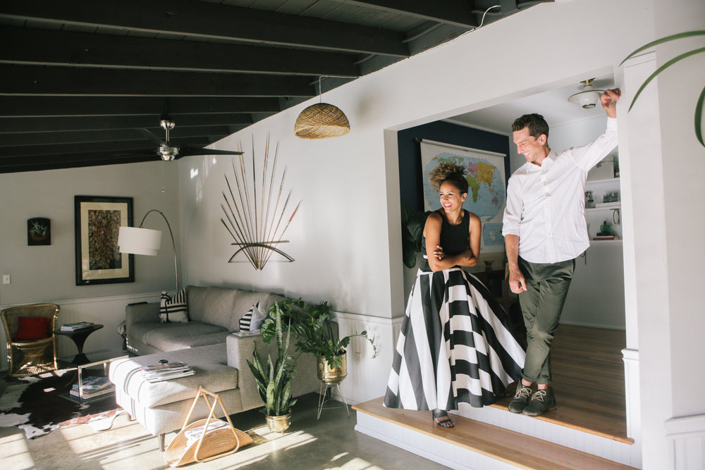 A woman in a black and white striped skirt and a man in a white shirt and green pants stand smiling in a stylish living room with modern decor and plants.