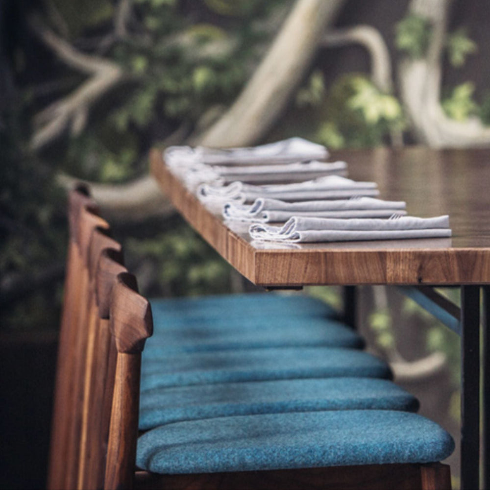 The WOOD High Stool (Bar Height) by Earl Home is a handcrafted wooden bar stool made in walnut, white oak, or ebonized oak, a hand shaped hand crafted backrest, footrest, and simple design. It offers customizable upholstery to match any decor. These stools as seen in Otium, Pendry Hotel, Provisional.