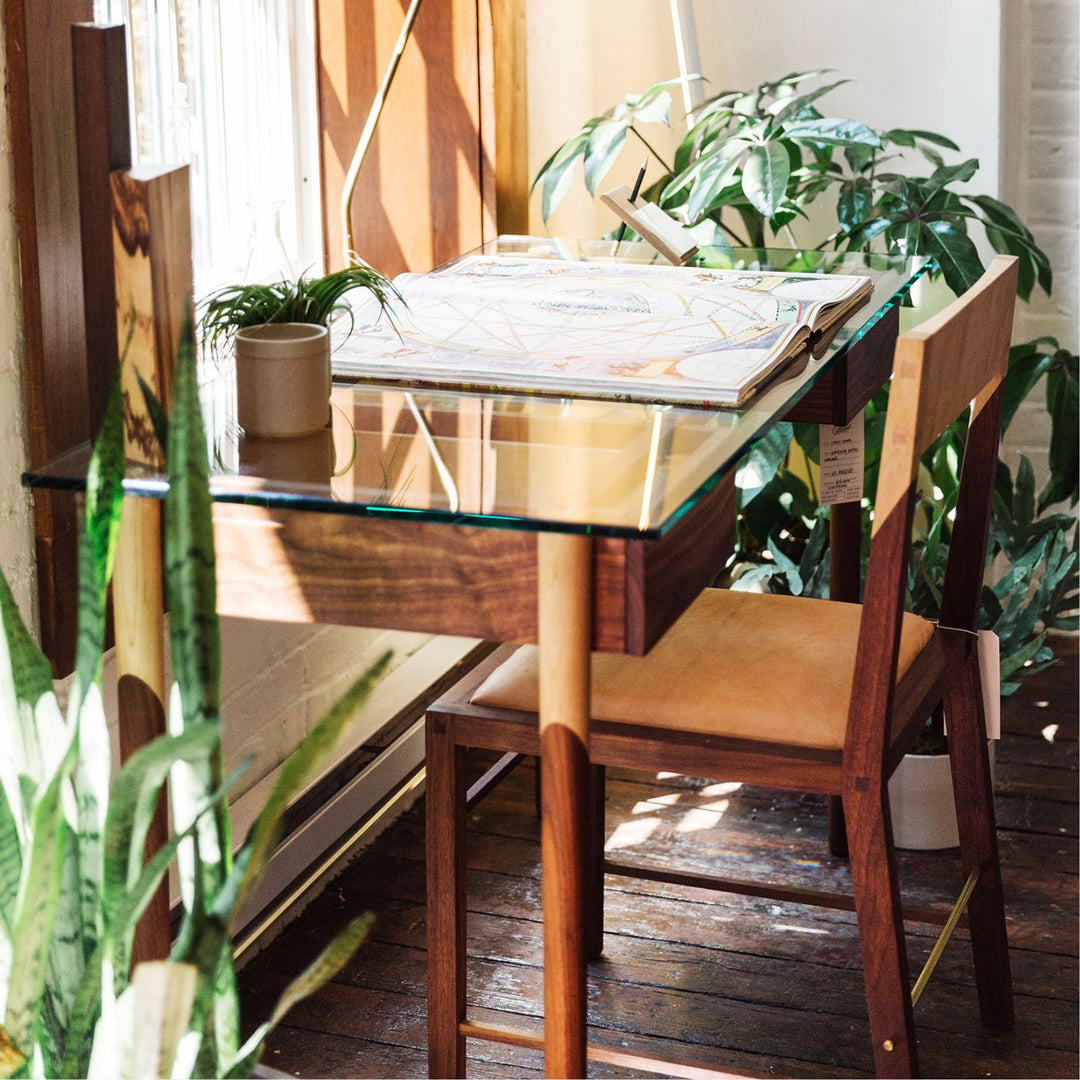 The WRITING Desk by Earl Home is an elegant glass desk with walnut and maple spliced wooden legs and a tempered glass top, Accompanied by a matching wooden chair. You can customize it to create your perfect workspace.