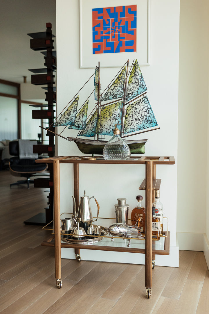 The BAR Cart from Earl Home is a solid wood bar cart on wheels with exquisite hand-cut joinery, featuring two shelves: a robust wood top and an elegant lower shelf made of tempered glass panels.