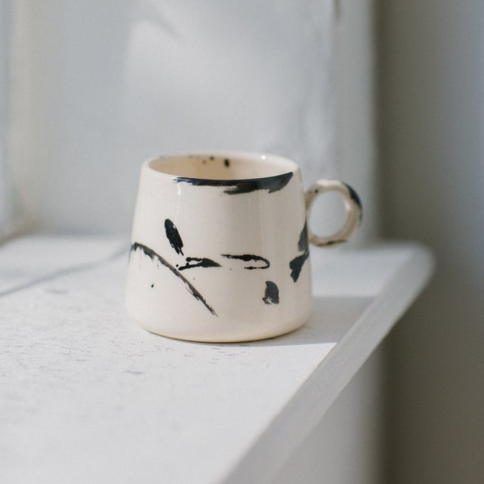 The TAPERED Mug by Earl Home is a hand-thrown white ceramic piece, showcasing a bold dark blue brush stroke and a round handle against a light gray background.