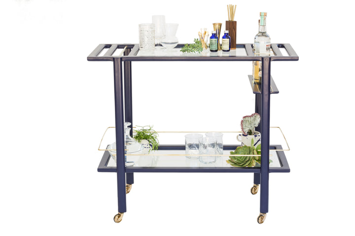 The BAR Cart from Earl Home is a solid wood bar cart on wheels with exquisite hand-cut joinery, featuring two shelves: a robust wood top and an elegant lower shelf made of tempered glass panels.